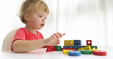 Child destroys a built house. Charming infant boy sitting at table and playing with colorful constructor blocks on white background