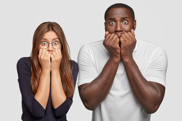Photo of astonished puzzled two interracial man and woman tremble from great fear, bite finger nails, have fearful expressions, stand closely to each other against white background. Omg concept