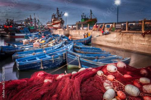 Plakat Maroko Essaouira portu w nocy. Selekcyjna ostrość na pierwszoplanowej czerwonej ryba kompensaci i pławikach. Tło ruchu plama kolorowe błękitne łodzie i seagulls w locie. Malownicza wioska rybacka.