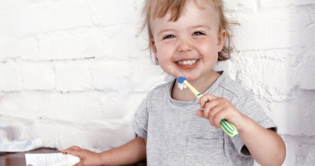Cute small blond boy in a gray shirt brushing his teeth with a toothbrush in bathroom