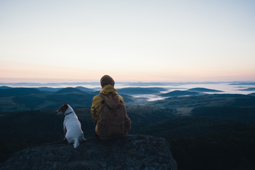Alone tourist sitting on the edge of the cliff with dog against the backdrop of an incredible mountain landscape. Sunny day and blue sky