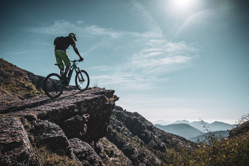 mountain biker riding up a large rock deep in the alps with sun and mountain layers behind