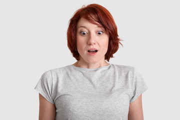 Scared red haired woman gazes in stupor on floor, dressed in grey t shirt, models over white background, cant believe in what she sees, isolated over white background. Feminity and reaction concept