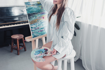 cropped view of artist sitting on chair in front of easel and holding palette in living room