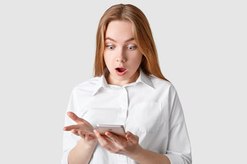 Shocked terrible woman stares at screen of smart phone, reads shocking news, dressed in elegant shirt, isolated over white background. Indoor shot of stupefied girlfriend with modern gadget.