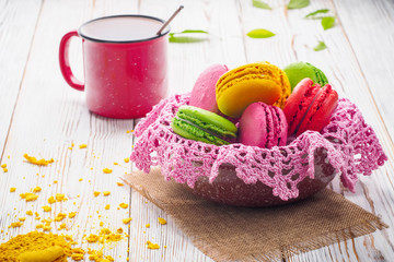 Assorted colorful sweet gentle soft French macaroons dessert cake macarons biscuit in bowl with knitted pink napkin on bright white table with red mug of tea and crumbled cake.
