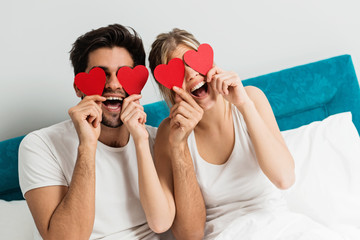 Young happy couple sitting on the bed, holding red hearts 