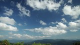 White clouds float on the blue summer sky. Below is a panorama of the mountain landscape. Time-Lapse Motion 4K video.