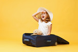 A little blonde child girl dreams of traveling and sits in a suitcase with a toy bear.Yellow background