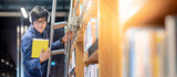 Asian man university student choosing book from bookshelf while standing on the ladder in library for education research. Bestseller collection in bookstore. Scholarship or educational opportunity