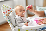 family, food, eating and people concept - father feeding little baby sitting in highchair with puree by spoon at home