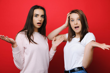 Two amazing girl friends looking into camera amazed with open mouths gesticulates with their hands on a red background.