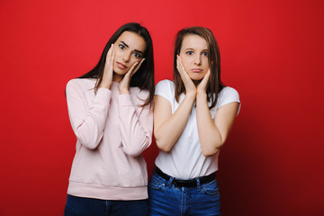 Two pretty female looking into camera astonished while holding her faces with both hands against a red wall.
