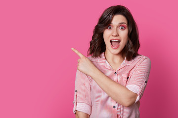 Indoor shot of surprised young woman with opened mouth, points with fore finger on blank space, shows incredible thing, wears striped shirt, isolated over pink background. Advertisement concept