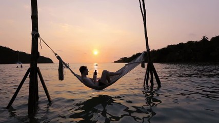 Man using smartphone on the beach hammock at sunset, mobile phone and travel.