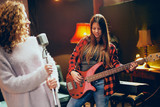 Two women practicing for the gig. Woman with curly hair holding microphone and singing while other one playing bass guitar. Home studio interior.