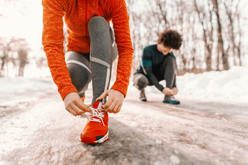 Close up of woman tying shoelace while crouching on the trail at winter. Outdoor fitness concept. In background man..