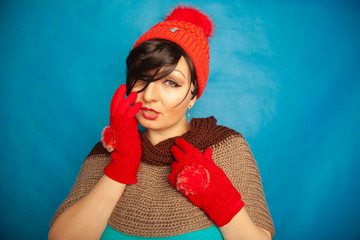 pretty brunette girl wearing fashion red knitted winter gat and warm beautiful gloves on blue studio solid background