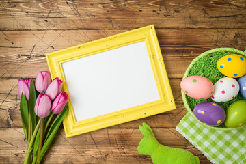 Easter holiday background with photo frame; easter eggs in basket and tulip flowers on wooden table.