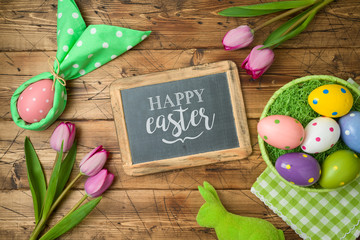 Easter holiday background with chalkboard, easter eggs in basket and tulip flowers on wooden table.