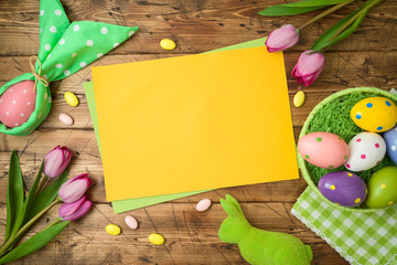 Easter holiday background with paper note, easter eggs in basket and tulip flowers on wooden table.