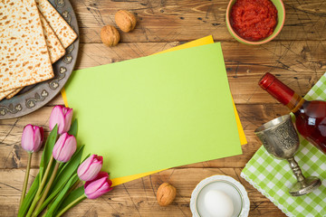 Jewish holiday Passover background with matzo, seder plate, wine, tulip flowers and paper note on wooden table.