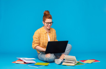 education, high school, technology and people concept - red haired teenage student girl in checkered shirt and torn jeans with books using laptop computer over bright blue background