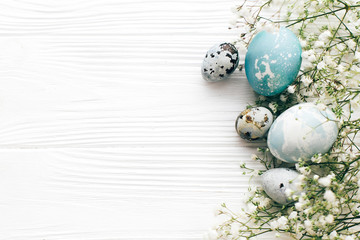 Happy Easter. Stylish Easter eggs with spring flowers border, flat lay on white wooden background with space for text. Modern easter eggs painted with natural dye in blue and grey marble.