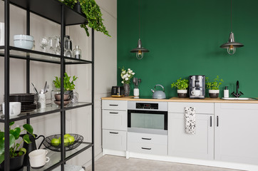 Two industrial lamps above kitchen furniture with herbs, coffee maker and roses in vase, copy space on empty green wall