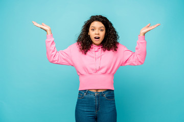 surprised curly african american woman showing shrug gesture isolated on blue