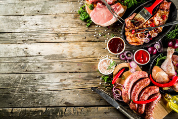 Various raw meat ready for grill and bbq, with vegetables, greens, sauces kitchen grilling utensils. Chicken legs, pork steaks, sausages, beef ribs with herbs, rustic wooden background copy space