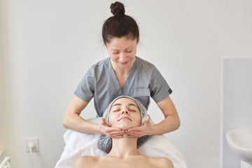 Cosmetologist wears gray gown massaging patient face. Young woman with naked shoulders and dresses madical cap has beauty facial procedures in salon. Healthcare, clinic, cosmetology concept.