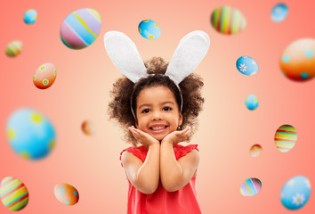easter, holidays and childhood concept - happy little african american girl wearing bunny ears headband posing over living coral background and colored eggs