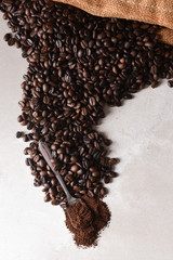 High angle view of a burlap bag of roasted coffee beans spilling onto the surface with a spoonful of coffee grounds