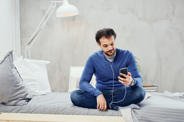 Happy cute arab man sitting on the bed in bedroom with legs crossed and listening music over smart phone.
