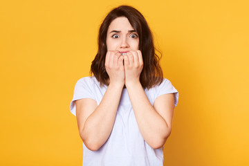 Studio shot of young brunette woman wearing casually, nervous and bites nails, standing with eyes full of fear, attractive female afraids of something, photo isolated on concentrated yellow background