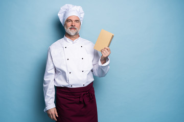 Portrait of a professional chef in uniform holding recipe book and looking at camera on light blue.
