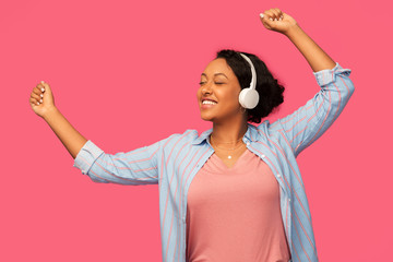 people, technology and audio equipment concept - happy african american young woman in headphones listening to music and dancing over pink background