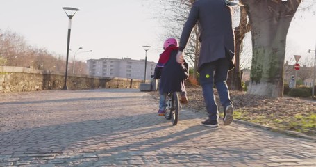 Daughter child girl learning riding bycicle with dad teaching in city.Growing,childhood,active safety family.Sidewalk urban outdoor.Warm sunset cold weather backlight.4k slow motion 60p back video