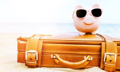 Pink piggy bank in sunglasses on leather travel case over sea beach background. Savings for vacation concept