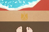 Feet on the sunny sandy beach with flag Egypt. View from top on surf.