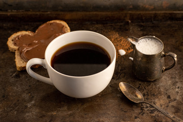 three-quarter view of black coffee with creamer and toast with chocolate in rustic dark setting