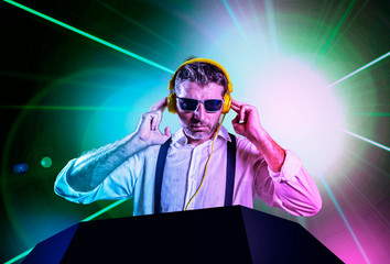 young attractive and cool DJ in shirt and suspenders remixing music at night club using headphones in party strobo and laser lights background in clubbing and nightlife