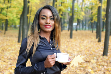 Beautiful young African woman drinking coffee from a white Cup on the background of autumn leaves in the Park