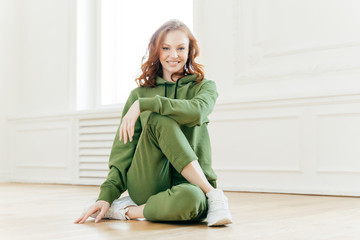 Positive redhead fitness girl feels relaxed, wears tracksuit, sportshoes, has rest after yoga training indoor, poses on floor, engaged in fitness, stays flexible, gets fit. Pilates exercising.