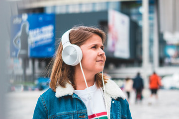 Cute lady wearing a blue jeans jacket listening to music on a big white pair of headsets in an urban city scape – Smiling young woman expressing joy and looking up while listening music