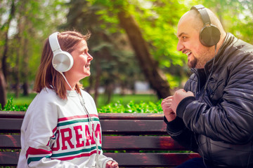Boy wearing a black leather jacket and girl with a white blouse enjoying music at their big headphones – Young man and woman sitting on a bench outdoor in nature looking at each other