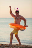 A man, wearing a rubber ring and a rim with horns, dances at the seashore at sunset