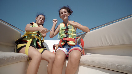 Two happy teen girls wearing safety belts rise hand up in the air and have fun on ride with luxury yacht
