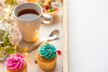 Bright cakes. Cupcakes with a cup of coffee or tea and color dots on a tray with a flowering branch and luminous garland on a white background